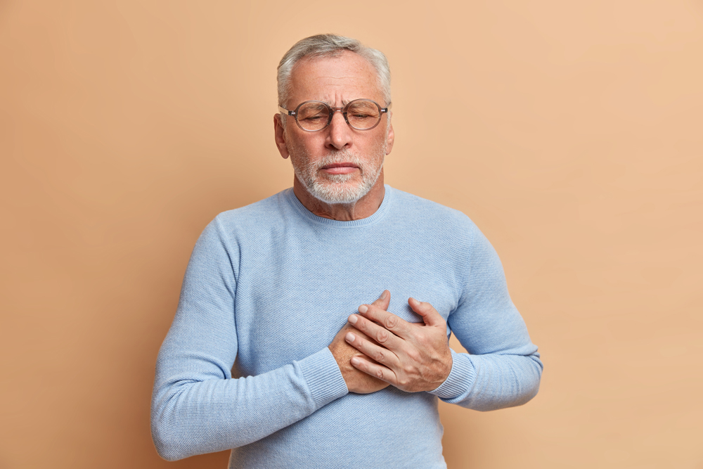 People age and health problems concept. Grey haired displeased bearded old man has sudden painful spasm in chest closes eyes and presses hands to heart poses against beige wall. Risk of heart attack